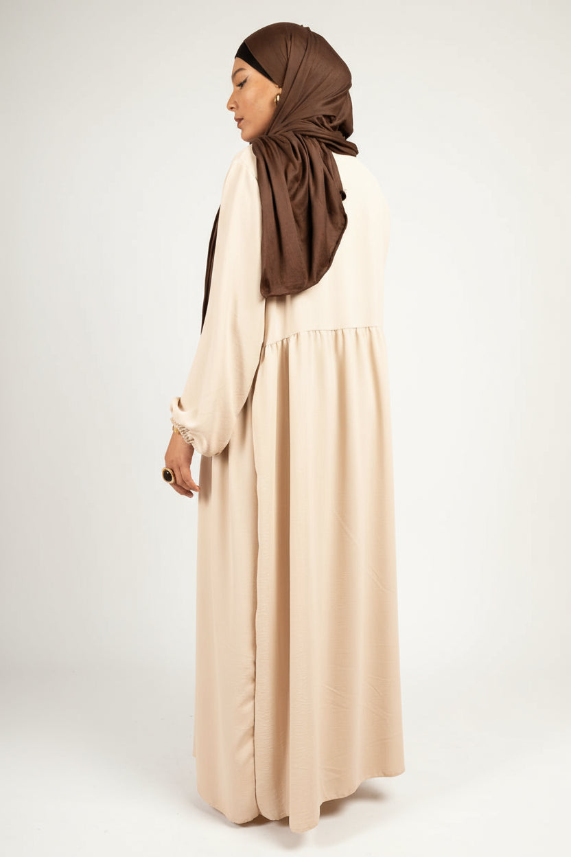 Woman wearing a long beige dress and brown hijab on a pink background