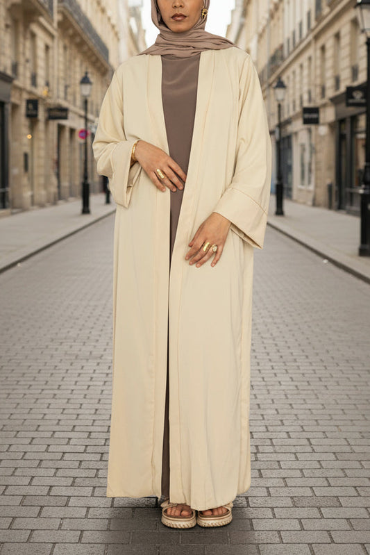 Woman wearing a long beige abaya over a brown dress on a pink background